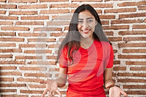 Young teenager girl standing over bricks wall smiling cheerful with open arms as friendly welcome, positive and confident