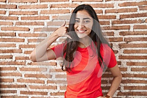 Young teenager girl standing over bricks wall smiling doing phone gesture with hand and fingers like talking on the telephone