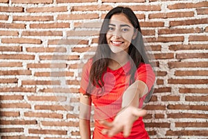 Young teenager girl standing over bricks wall smiling friendly offering handshake as greeting and welcoming