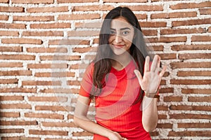 Young teenager girl standing over bricks wall waiving saying hello happy and smiling, friendly welcome gesture