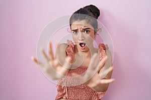 Young teenager girl standing over pink background afraid and terrified with fear expression stop gesture with hands, shouting in