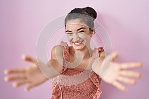 Young teenager girl standing over pink background looking at the camera smiling with open arms for hug