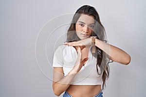 Young teenager girl standing over white background doing time out gesture with hands, frustrated and serious face