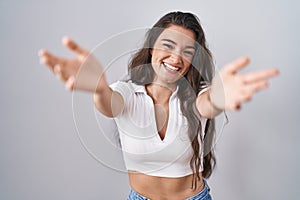 Young teenager girl standing over white background looking at the camera smiling with open arms for hug