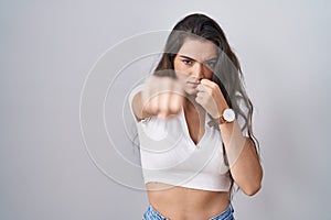 Young teenager girl standing over white background punching fist to fight, aggressive and angry attack, threat and violence