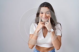 Young teenager girl standing over white background smiling with open mouth, fingers pointing and forcing cheerful smile