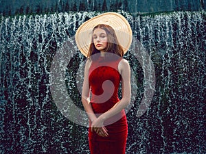 Young teenager girl in stylish red dress in unique location in water and water fall in the background. Lady in high fashion outfit