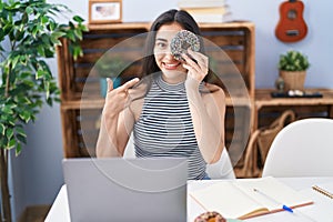 Young teenager girl using computer laptop with doughnut on face smiling happy pointing with hand and finger