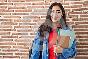 Young teenager girl wearing student backpack and holding books looking positive and happy standing and smiling with a confident