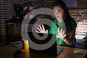 Young teenager girl working at the office at night afraid and terrified with fear expression stop gesture with hands, shouting in