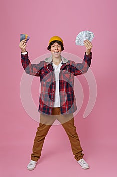 Young teenager guy 17s hold credit, debit cards and money in hands waring hat and plaid jacket isolated on pink
