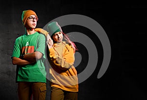 Young teens boy and girl in green and yellow comfortable casual clothing and hats standing and looking aside over dark background