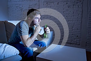 Young television addict man sitting on home sofa watching TV and drinking beer bottle