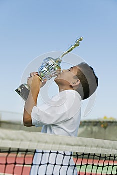 Young tennis player by net on court kissing trophy side view