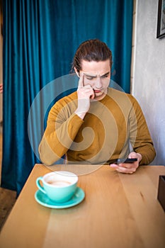 Young thinking man using phone and holding coffee cup