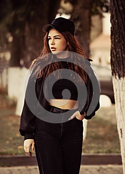 Young thinking teen hipster woman looking near the tree  on the street background in black style clothing and cool cap. Autumn