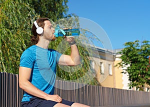 Young thirsty sportsman drinking fresh water from bottle while running outdoors on sunny day