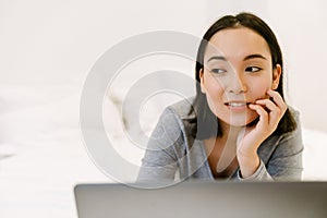 Young thoughful asian girl lying with laptop on bed