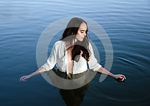 Young thoughtful brunette woman in white shirt is standing in the lake and moving hands through water.