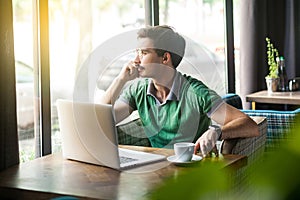 Young thoughtful businessman in green t-shirt sitting and working on laptop, looking to outside and thinking about his plans