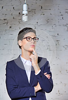 Young thoughtful businesswoman looking at energy efficient lightbulb hanging against brick wall at office