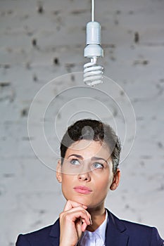 Young thoughtful businesswoman looking at energy efficient lightbulb hanging against brick wall at office