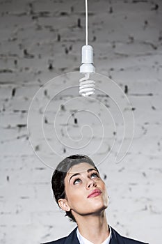 Young thoughtful businesswoman looking at energy efficient lightbulb hanging against brick wall at office