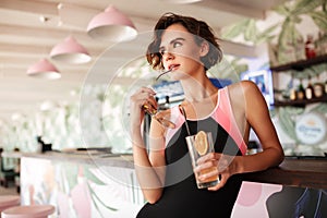 Young thoughtful girl in trendy swimsuit leaning on beach bar counter with sunglasses and cocktail in hands. Portrait of