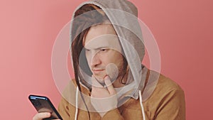 Young thoughtful man browsing mobile phone on pink background. Pensive male using smartphone and scratching chin.