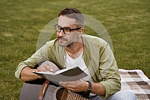 Young thoughtful man wearing eyeglasses holding acoustic guitar and composing a song while sitting on a green grass
