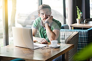 Young thoughtful quizzical businessman in green t-shirt sitting with laptop, hand on chin looking away and thinking what to do