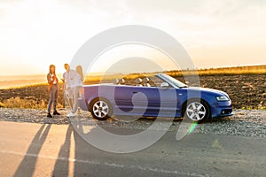 Young three friends standing near convertible car and talking each other have fun on beauty sunset on the road