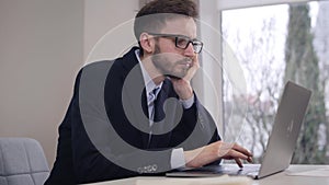 Young tired Caucasian businessman holding head with hand and typing on laptop keyboard. Bored handsome man taking off