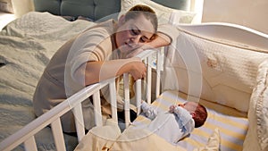 Young tired and exhausted mother fallen asleep while rocking crib of her newborn baby at night. Concept of sleepless