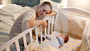 Young tired and exhausted mother fallen asleep while rocking crib of her newborn baby at night. Concept of sleepless