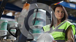 Young tired female forklift driver taking off hard hat sighing sitting in autoloader in industrial warehouse indoors