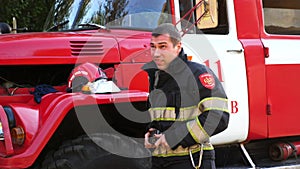 Young tired fireman unzipping protective workwear after hard work near a fire engine. Male firefighter after fire