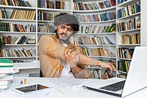 Young tired guy stretching hand and shoulder while looking at notebook on desk with books and computer. Bored male