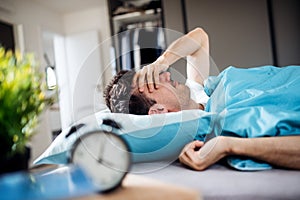 Young tired man with alarm clock in bed at home, waking up in morning.