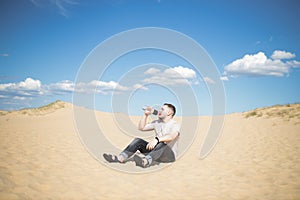 Young tired man drinking water while sitting on the top of sand dune in desert, sand is al around and some greens. Hot summer weat