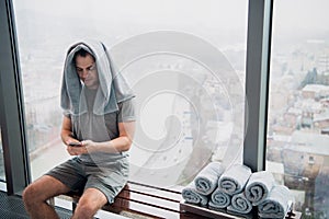 Young, tired man sitting on bench in a gym. Holding mobile phone and towel on his head. Gym at skyscraper with big