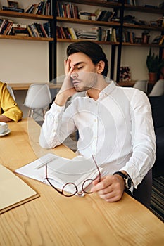 Young tired man sitting at the table with notepad leaning head on hand sleeping at work in modern office