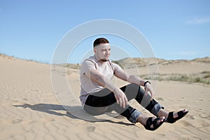 Young tired man sitting on the top of sand dune in desert, sand is al around and some greens. Hot summer weather, concept of trave