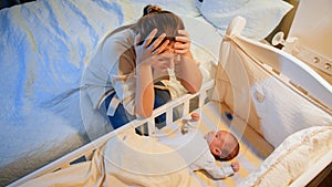 Young tired mother suffering from depression lookin on her sleepless newborn baby in crib at night. Maternal depression photo