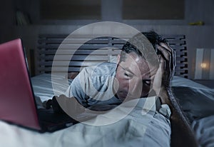 Young tired and overworked man lying and networking on bed at home bedroom sleepy working with laptop computer late at night