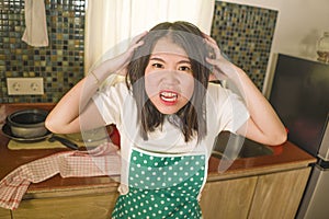 Young tired and stressed Asian Korean woman in cook apron working in the kitchen screaming desperate and pulling her hair