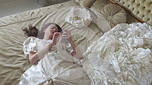 Young tired woman in ball gown lying on the gold decorated bed and texting on cell phone close up.