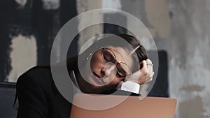 Young tired woman in glasses sitting at the laptop computer while working in the office, then almost falling asleep and