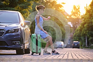 Young tired woman with suitcase sitting beside car waiting for someone. Travelling and vacations concept