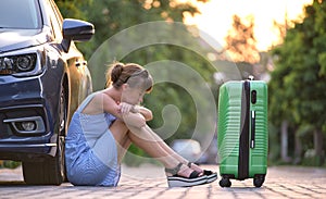 Young tired woman with suitcase sitting near her car waiting for someone. Travel and vacations concept